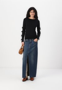 Schwarzes Langarmoberteil mit Schleifenakzenten an den Ärmeln, kombiniert mit einem blauen Jeans-Maxirock mit einem Schlitz an der Vorderseite. Braune Handtasche und Blockabsatzstiefel.