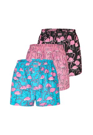 Muskelbepackter Mann mit verschränkten Armen trägt pinke Shorts mit Flamingo-Muster und steht vor einem einfarbigen hellgrauen Hintergrund.