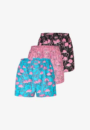 Muskelbepackter Mann mit verschränkten Armen trägt pinke Shorts mit Flamingo-Muster und steht vor einem einfarbigen hellgrauen Hintergrund.
