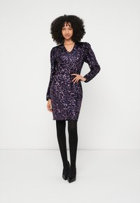 Fialové šaty s flitry, s V-výstřihem, dlouhými nafouklými rukávy a přiléhavým střihem, doplněné černými punčocháči a špičatými podpatky.