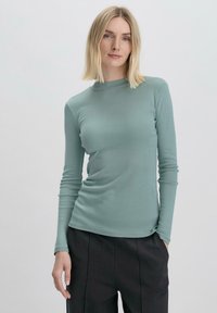 Maglietta a maniche lunghe in morbido tessuto verde chiaro con un collo alto e un design aderente, abbinata a pantaloni scuri e larghi.