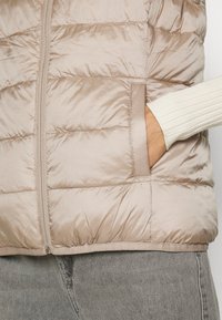 Chaleco acolchado color beige claro con textura acolchada, cierre frontal con cremallera y bolsillo lateral; combinado con una manga blanca acanalada.