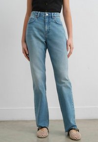 Jeans de cintura alta de color azul claro con un diseño de pierna recta, que cuentan con bolsillos frontales y un cierre de un solo botón. Llevados con zapatos de malla beige.