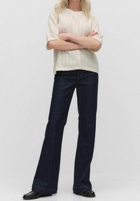 Krémový pletený cardigan s krátkým rukávem a strukturálními vzory, doplněný o tmavě modré rozšířené džíny a černou obuv.