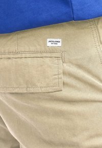 Pantaloni cargo beige in tessuto con una tasca posteriore e un'etichetta bianca ricamata che dice "JACK & JONES EST. 1990." Parte superiore blu navy parzialmente visibile.