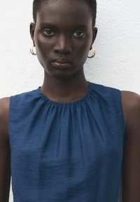 Námornícky modrý bezrukávový top s nariaseným výstrihom; vyrobený z jemnej látky. Model má na sebe veľké zlaté kruhové náušnice na textúrovanom bielom pozadí.