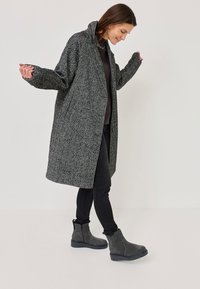 Harmaa, teksturoitu pitkä takki korkealla kauluksella, jossa on sivutaskut ja rento istuvuus, yhdistettynä tummiin slim-fit-farkkuihin ja harmaisiin nilkkureihin.