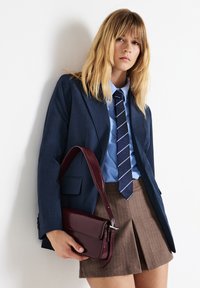 Blazer blu navy con risvolti appuntiti indossato sopra una camicia azzurro chiaro e una cravatta a righe, abbinato a una gonna pieghettata marrone. In mano una borsa a tracolla in pelle bordeaux.