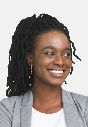 Donna con lunghe dreadlocks nere, che indossa orecchini a catena dorati, un blazer grigio chiaro e una maglietta bianca, sorridente su uno sfondo neutro.