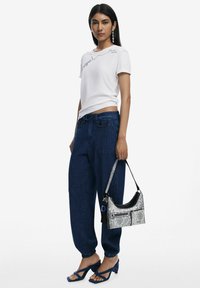 White T-shirt with embroidered detail, dark blue denim pants, and blue strappy heels. Silver snakeskin handbag with black accents.