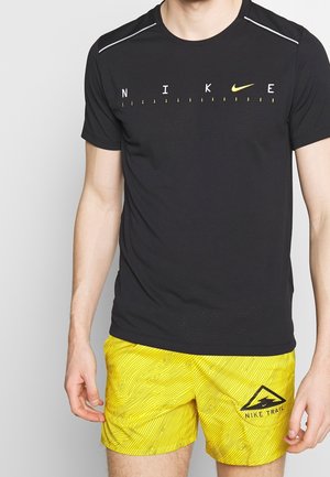 Mężczyzna w czarnej koszulce Nike z logo oraz żółtych krótkich spodenkach Nike Trail z topograficznym wzorem na jednolitym tle.