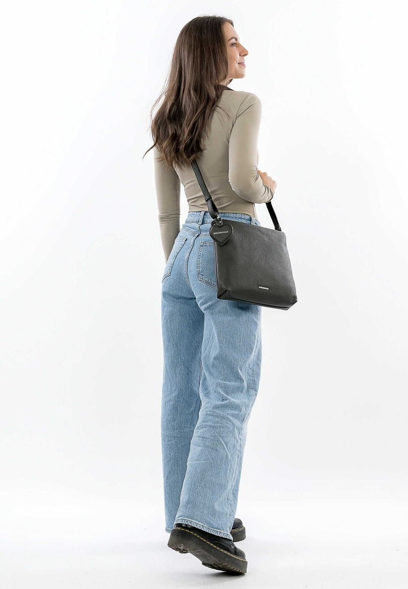 Black leather shoulder bag with textured surface, flat rectangular shape, long strap, and circular hardware detail. Worn with light blue jeans.