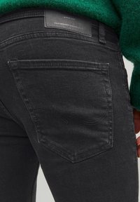 Jeans de mezclilla negra con un bolsillo trasero que presenta un detalle de costura curva. La tela tiene una ligera textura, con un parche de logo de cuero en la cintura.