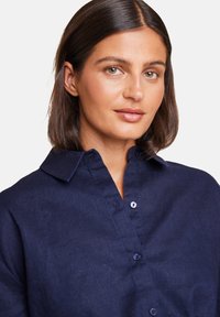 Marineblå lin skjorte med knappet krage, som har et teksturert stoff og synlig søm. Nærbilde av modellens overkropp.