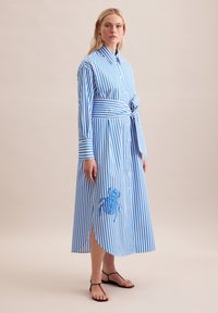 Abito a righe blu e bianche con bottoni, maniche lunghe, vita legata e uno spacco laterale. Presenta un dettaglio ricamato blu sulla gonna.