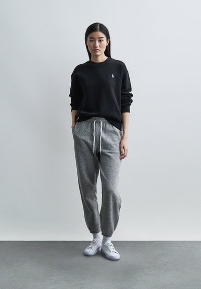 Γυναίκα στέκεται μπροστά σε γκρι τοίχο φορώντας μαύρο φούτερ, γκρι φόρμες με κορδόνι, λευκά αθλητικά παπούτσια, με τα χέρια στις τσέπες και ουδέτερη έκφραση.