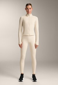 Light beige activewear set featuring a fitted long-sleeve zip jacket and matching leggings with a textured pattern, paired with black sneakers.