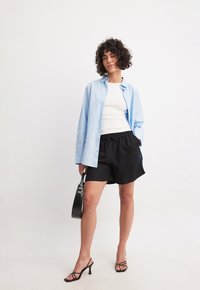 Camicetta blu chiaro, canotta bianca, pantaloni corti neri, borsa nera e sandali neri con cinturini. Capelli corti e ricci visibili.