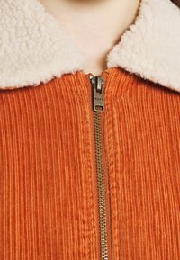 Veste en velours côtelé orange avec un col en fausse fourrure beige, dotée d'une fermeture éclair en métal et d'une texture côtelée.