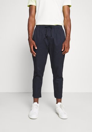 Homme debout portant un pantalon noir raccourci à cordon, des baskets blanches et une chemise blanche à manches courtes sur un fond uni.