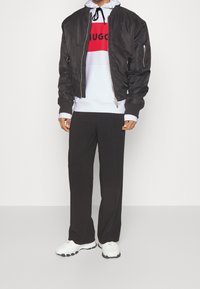 Schwarze Bomberjacke über einem weißen Hoodie mit rotem und schwarzem Logo, kombiniert mit lockeren schwarzen Hosen und weißen Sneakers mit schwarzen Sohlen.