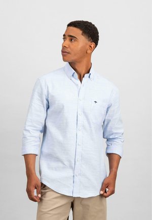 Hellblaues Button-up-Hemd aus strukturiertem Baumwollstoff; mit einer Brusttasche und umgeschlagenen Ärmeln; kombiniert mit beigen Hosen.
