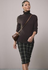 Pulover tricotat maro cu guler înalt, asortat cu o fustă creion în carouri negre și verzi. O geantă mică din răchită maro este aruncată pe umăr.