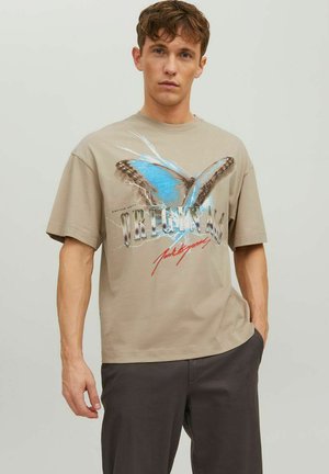 T-shirt grafico beige con una grande stampa di una farfalla blu con accenti marroni e la parola "ORIGINALE" in lettere metalliche e rosse.