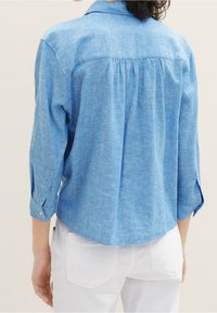 Chemise bleu clair à manches longues avec col, en tissu texturé, présentant un dos plissé et des manches retroussées, associée à un pantalon blanc.