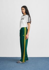Γυναίκα με μακριά σκουρόχρωμα μαλλιά στέκεται πλαγίως, φορώντας λευκό μπλουζάκι Adidas με μαύρες ρίγες και πράσινη φόρμα Adidas με κίτρινες ρίγες.