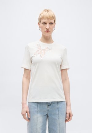 Femme aux cheveux blonds courts portant un haut blanc à manches courtes avec un détail en dentelle en forme de papillon et un jean taille haute bleu clair, debout devant un fond uni.