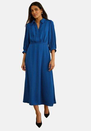 Kobieta w niebieskiej sukience midi z rękawami do połowy przedramienia, kołnierzykiem, pionowymi plisami oraz czarnymi szpilkami z zaokrąglonym noskiem, stojąca na tle jednolitej ściany.