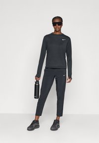 Svart langermet skjorte med en liten hvit Nike-logo, kombinert med svarte bukser og mørke sportssko. Modellen holder en svart vannflaske.