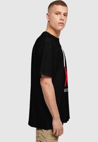 Sort oversized T-shirt lavet af bomuld, med et stort rødt grafisk motiv med ordet "X" og hvid tekst, korte ærmer og rund halsudskæring.