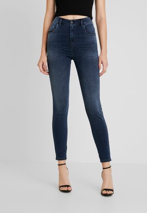 Femme portant un jean skinny taille haute bleu foncé et des talons noirs ouverts à l'avant avec une bride à la cheville, debout devant un fond clair uni.