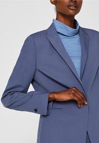 Blå kavaj med skarpa slag och knäppta manschetter, i kombination med en ljusblå polotröja under. Kavajen har en slät, skräddarsydd struktur.