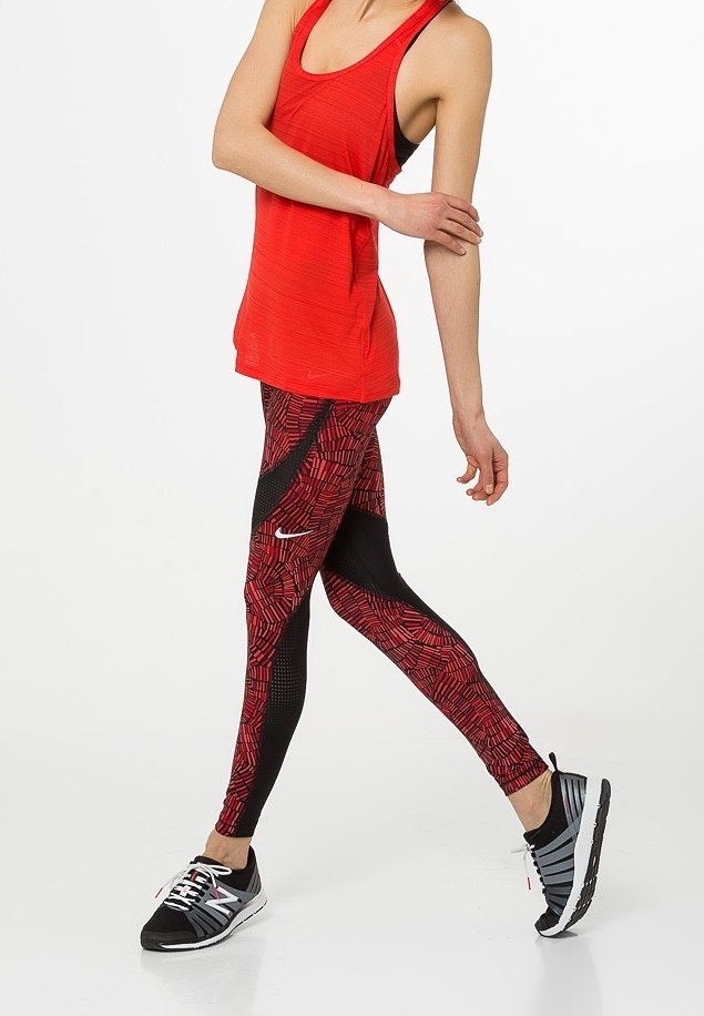 Femme marchant portant un haut rouge sans manches, des leggings rouges et noirs à motifs, et des chaussures de course noires et blanches sur fond blanc.
