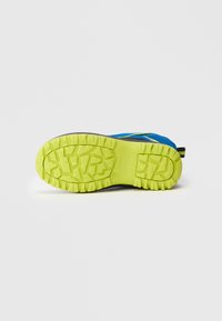 Blauwe en zwarte sneaker met een felgeel-groene zool die omhoog gericht is op een witte achtergrond.