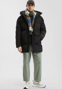 Čierna prešívaná bunda s kapucňou, ktorá má dve predné vrecká a zips. Model má na sebe pruhovaný pletený šál, svetlozelené nohavice a tenisky.