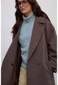 Przesadny brązowy wełniany płaszcz z dużymi wyłogami, nałożony na jasnoniebieski sweter w warkocze. Posiada duże, przyciemnione okulary przeciwsłoneczne i beżowe spodnie.