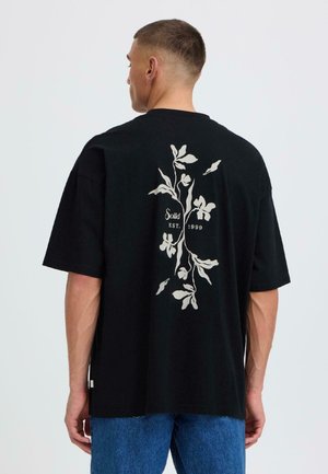 Homme portant un t-shirt noir oversize avec un motif floral blanc et le texte « Solid EST. 1999 » dans le dos, associé à un jean bleu.