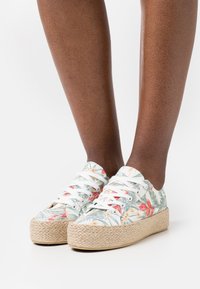 Scarpe di tela con motivo floreale e lacci bianchi, dotate di una spessa suola in juta intrecciata color beige. Il design include foglie e fiori tropicali.
