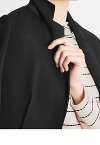 Blazer noir avec un col structuré et des lignes épurées, doté d'un tissu lisse et extensible. La chemise rayée en dessous a une texture légère et douce.