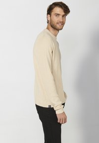 Beige ribbelshirt met lange mouwen en een ronde hals. De stof lijkt textuur te hebben, gecombineerd met zwarte aansluitende broek.