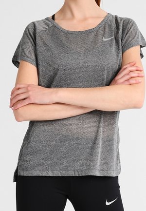 Mujer con camiseta gris texturizada de manga corta de Nike y leggings negros de Nike, con los brazos cruzados contra un fondo claro y liso.