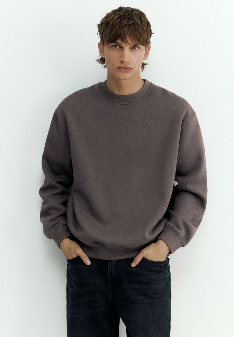 Jeune homme aux cheveux courts et bruns, portant un sweat-shirt marron uni à col rond et un jean noir, debout les mains dans les poches sur fond blanc.
