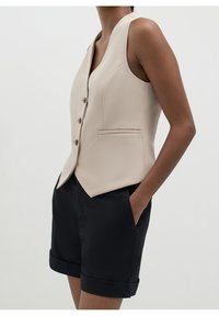 Gilet beige sans manches boutonné avec poches avant, associé à un short noir à revers, sur une personne debout avec une main dans la poche.