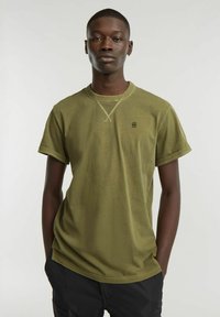 T-shirt verde oliva a maniche corte realizzato in tessuto morbido, con colletto a girocollo e un piccolo logo sul lato sinistro del petto. Design semplice e rilassato.