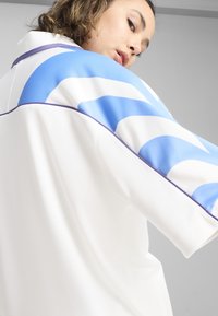 Haut de sport blanc avec des motifs de vagues bleus et blancs sur les manches, bord violet, tissu texturé et design ample.