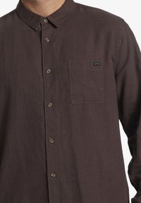 Brown linen long-sleeve shirt featuring a pointed collar, button closure, and a chest pocket with a small branded tag.